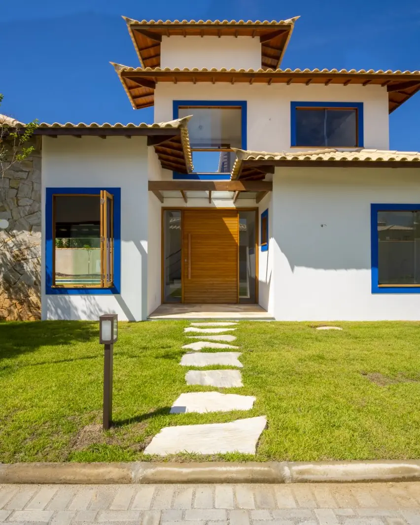 Fachada de uma residência tipo B, com porta de madeira, esquadrias azuis e pedra